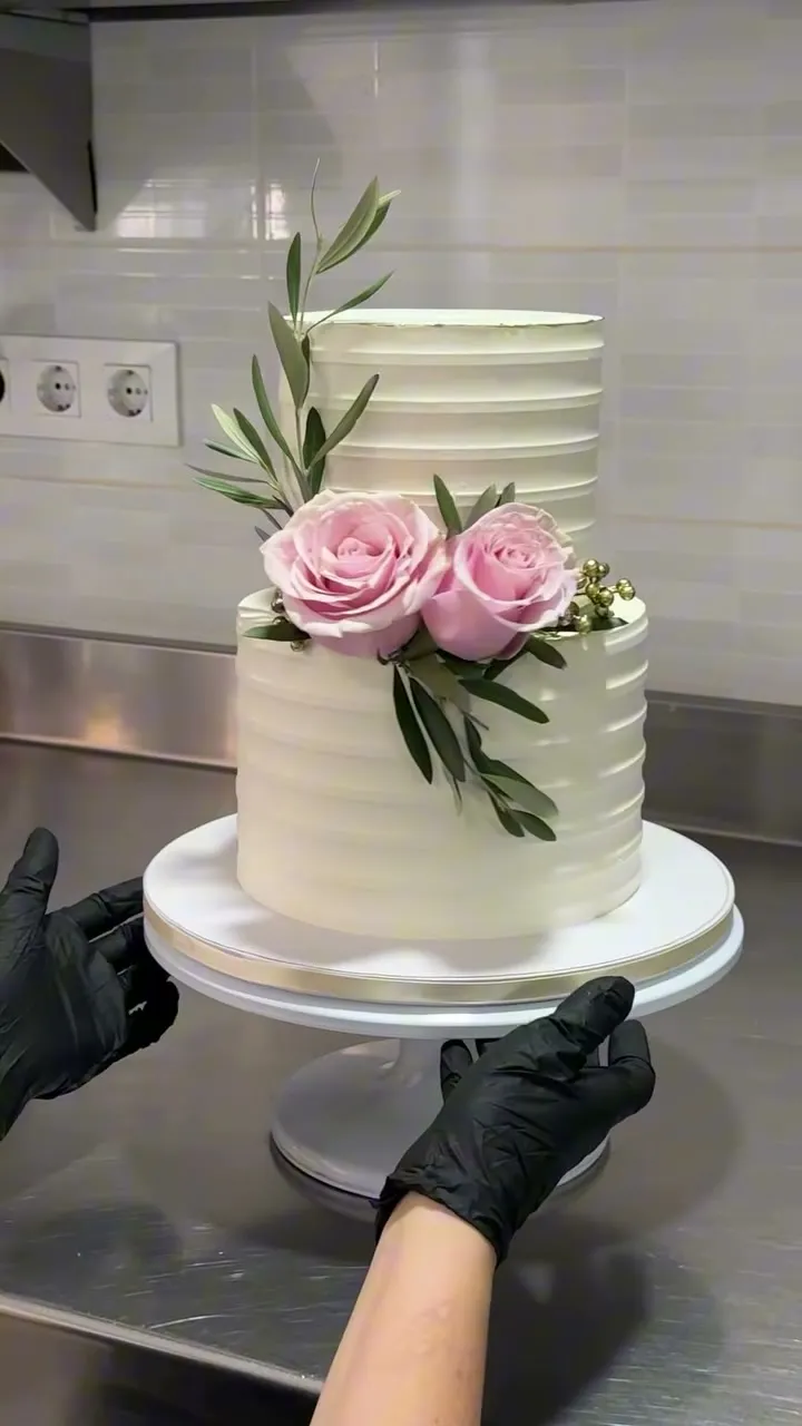 Tarta blanca de dos pisos con rosas y ramas de olivo, sostenida con guantes negros